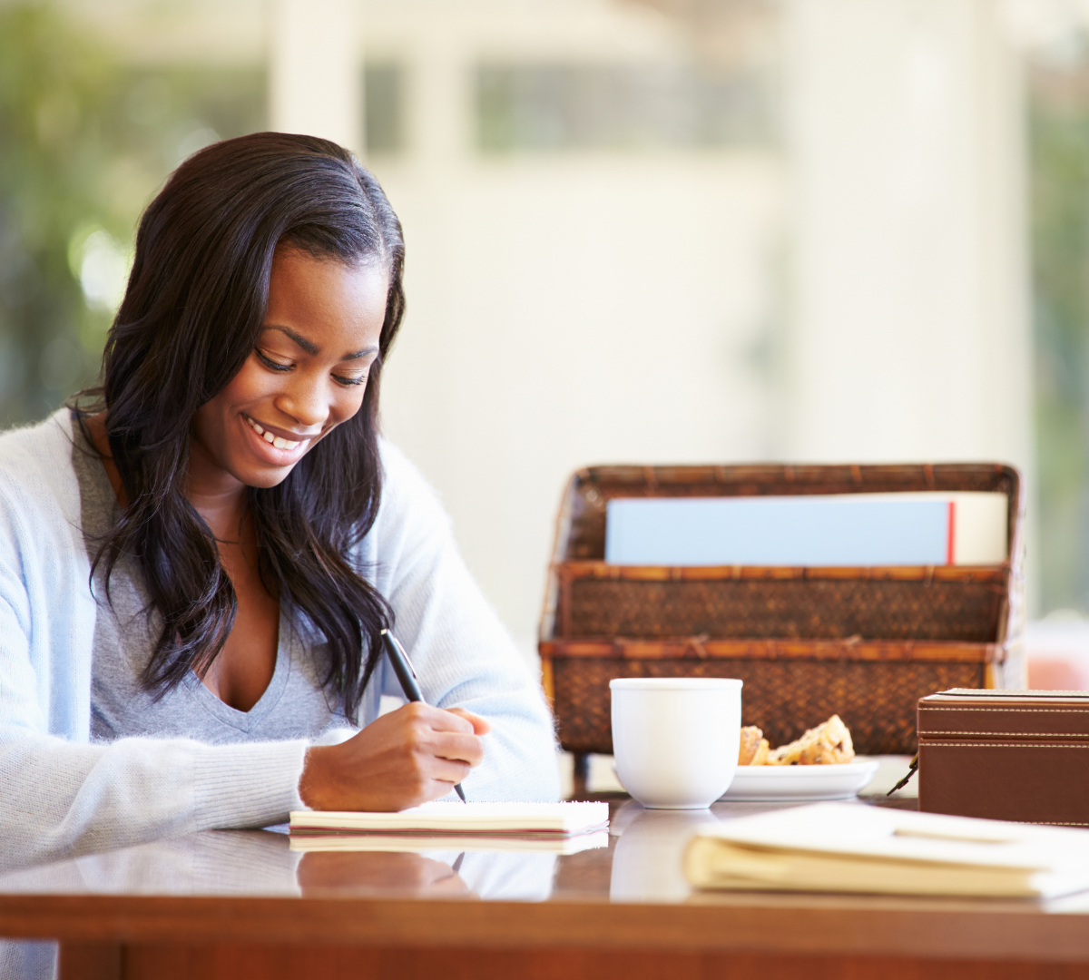 A black woman sitting at a desk and smiling. She holds a pen in her hand and is writing in a book